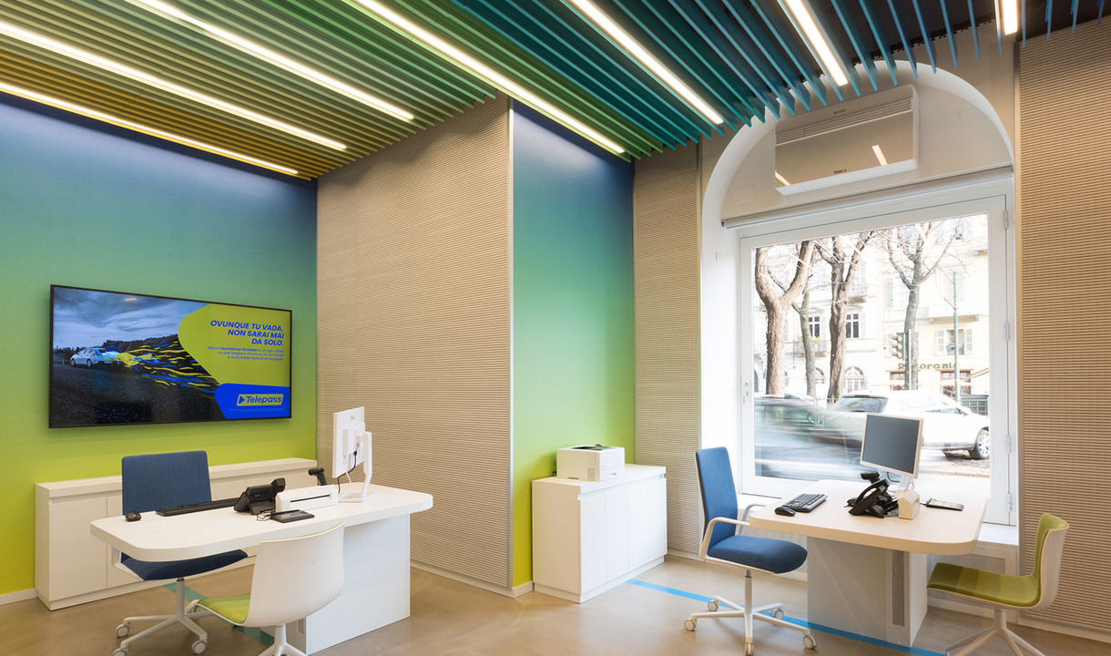 Modern office interior with Artemide linear ceiling lights, vibrant green-yellow gradient walls, white desks, and blue chairs.