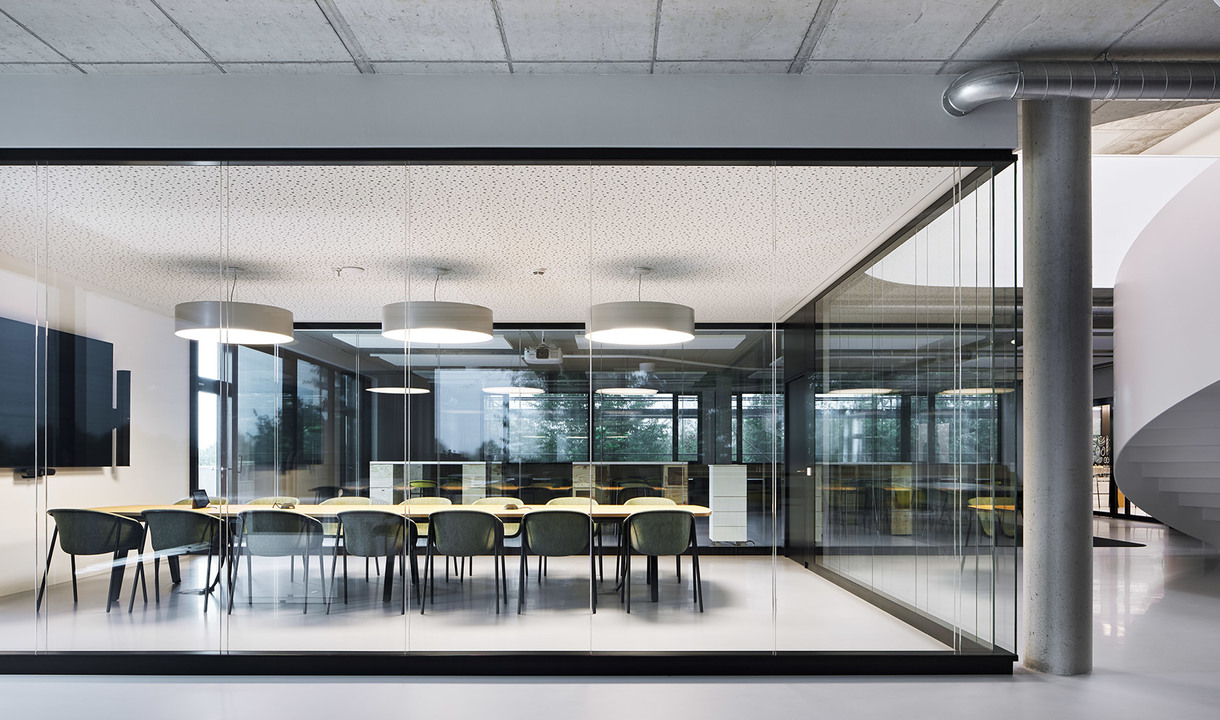 Modern glass-walled conference room with long table, green chairs, and two white cylindrical pendant lights overhead.