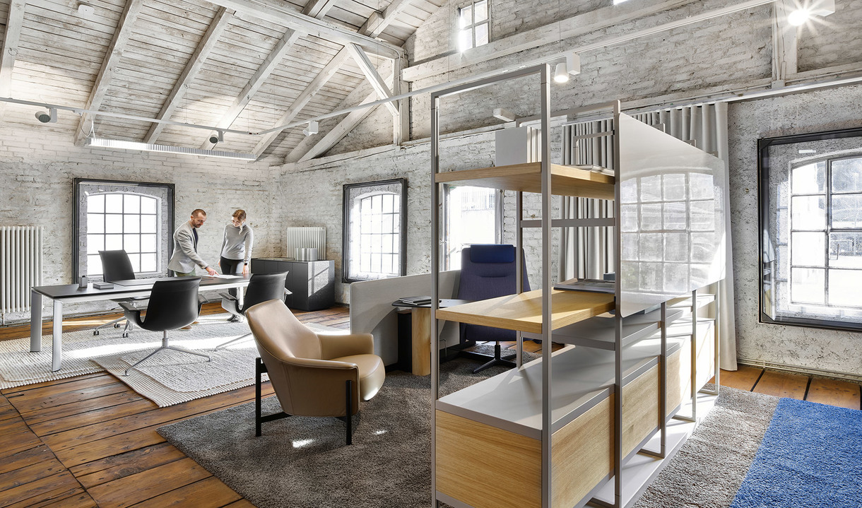Modern loft office with exposed brick walls, wooden beams, open shelving unit, leather armchair, and two people working at desks