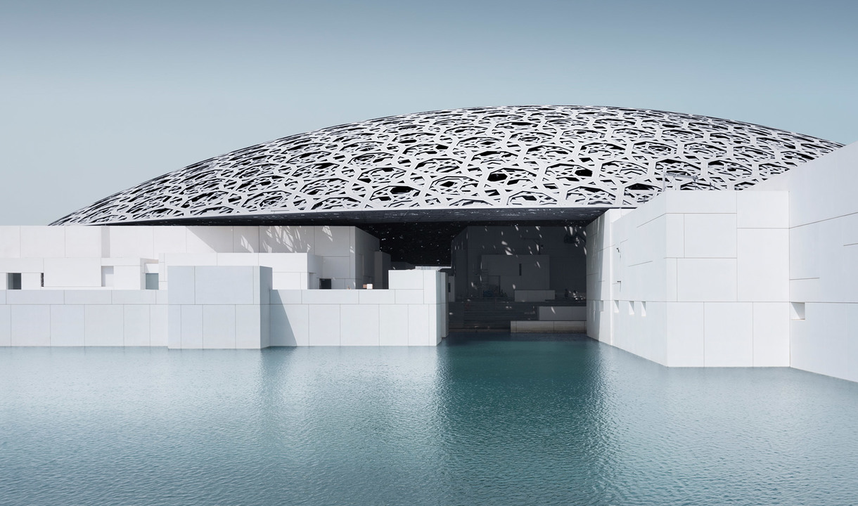 Louvre Abu Dhabi museum with iconic latticed dome reflecting on surrounding water, white geometric buildings under overcast sky
