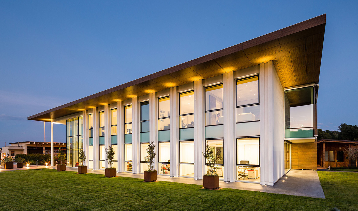 Modern two-story building with tall white columns and floor-to-ceiling windows, illuminated by warm architectural uplighting at dusk