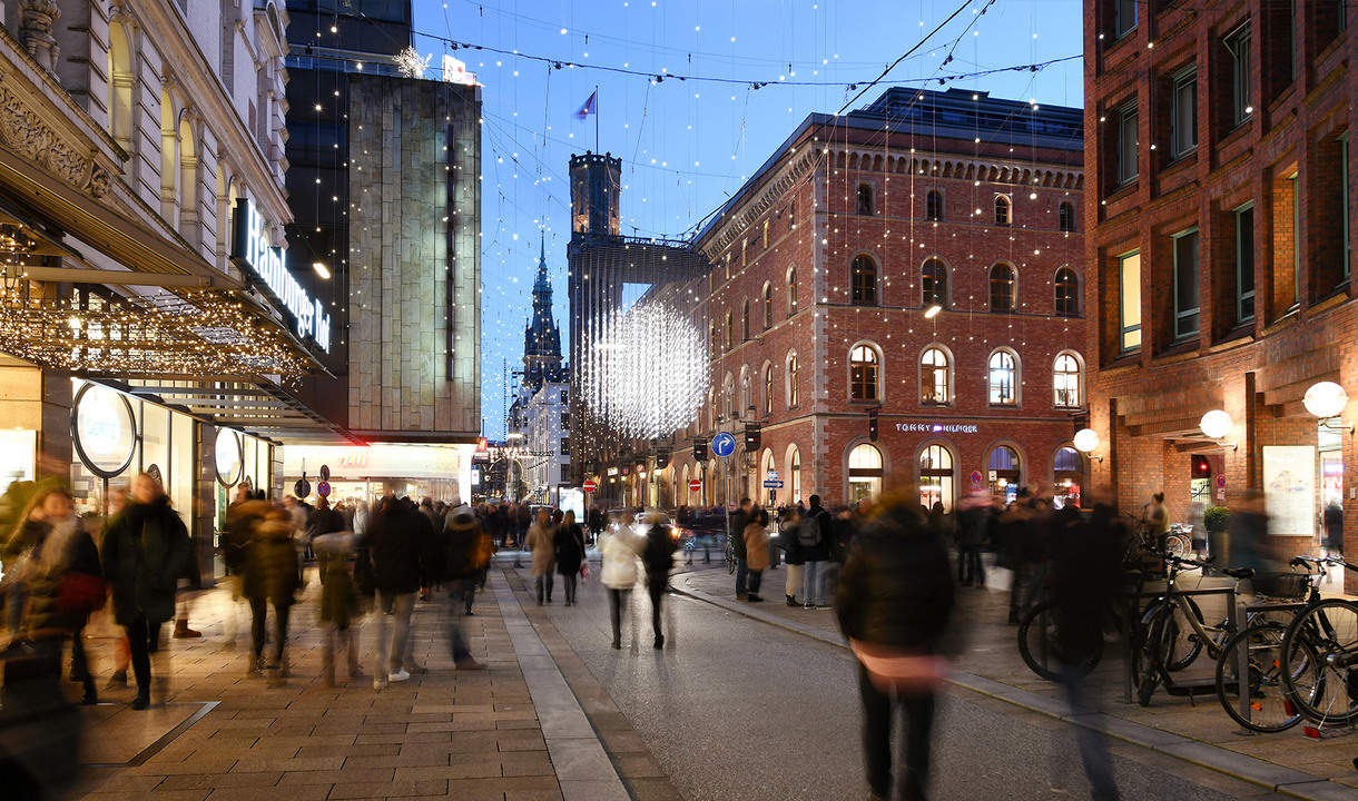 Busy pedestrian street at dusk with warm festive lights, glowing storefronts, and a large luminous installation above the crowd
