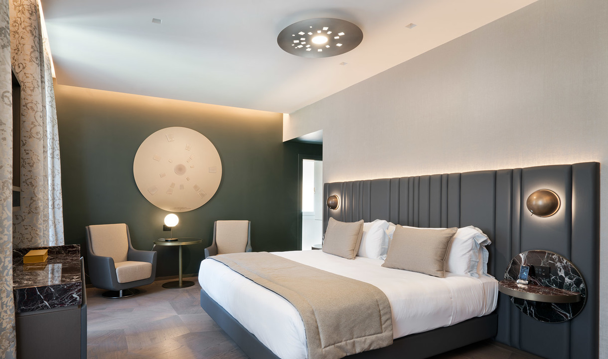 Elegant hotel room with Artemide ceiling, wall, and table lamps providing warm ambient lighting over a king-size bed and seating area