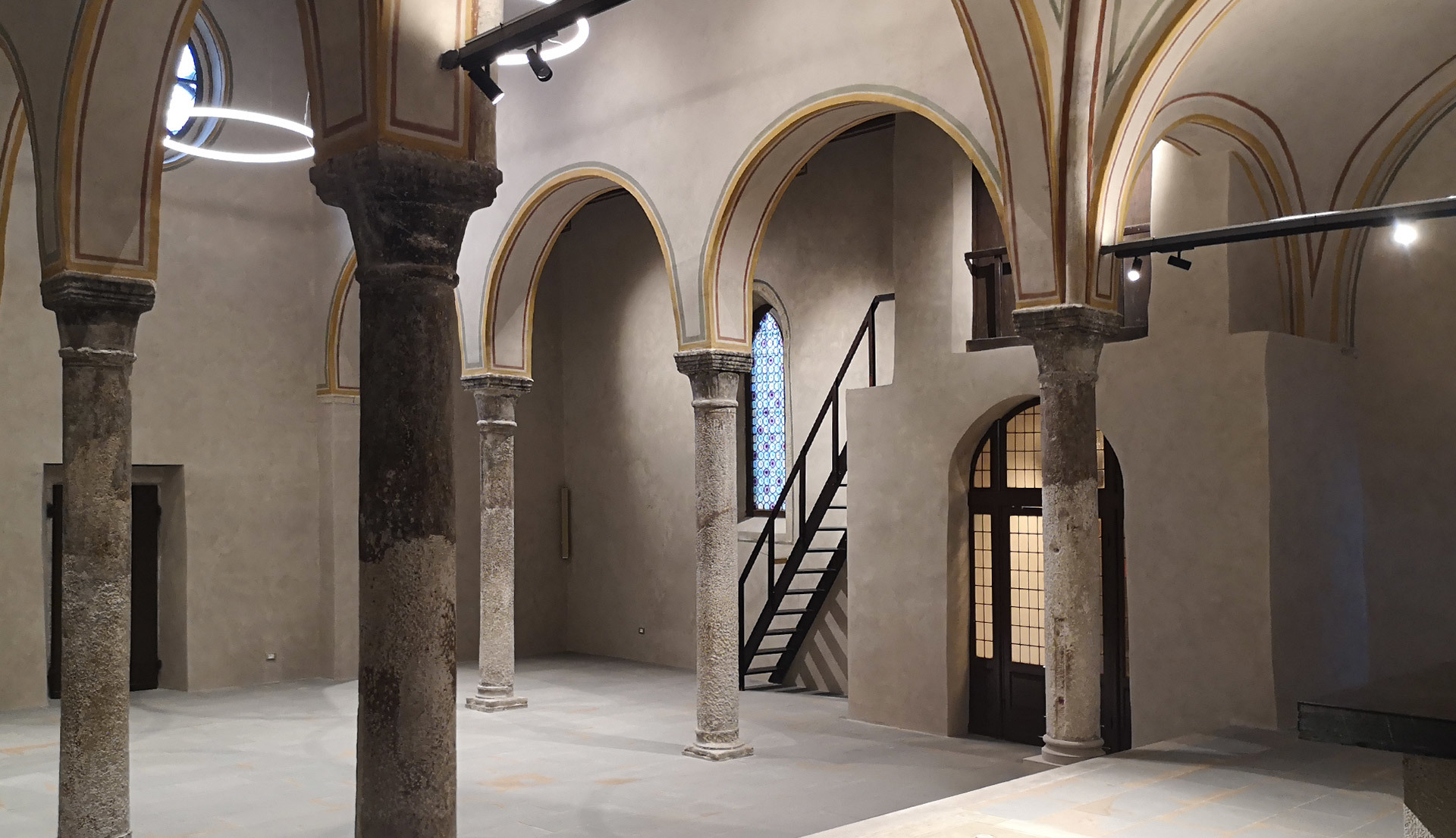Restored historic hall with stone arches and columns, illuminated by Artemide circular pendant and track spotlights overhead.