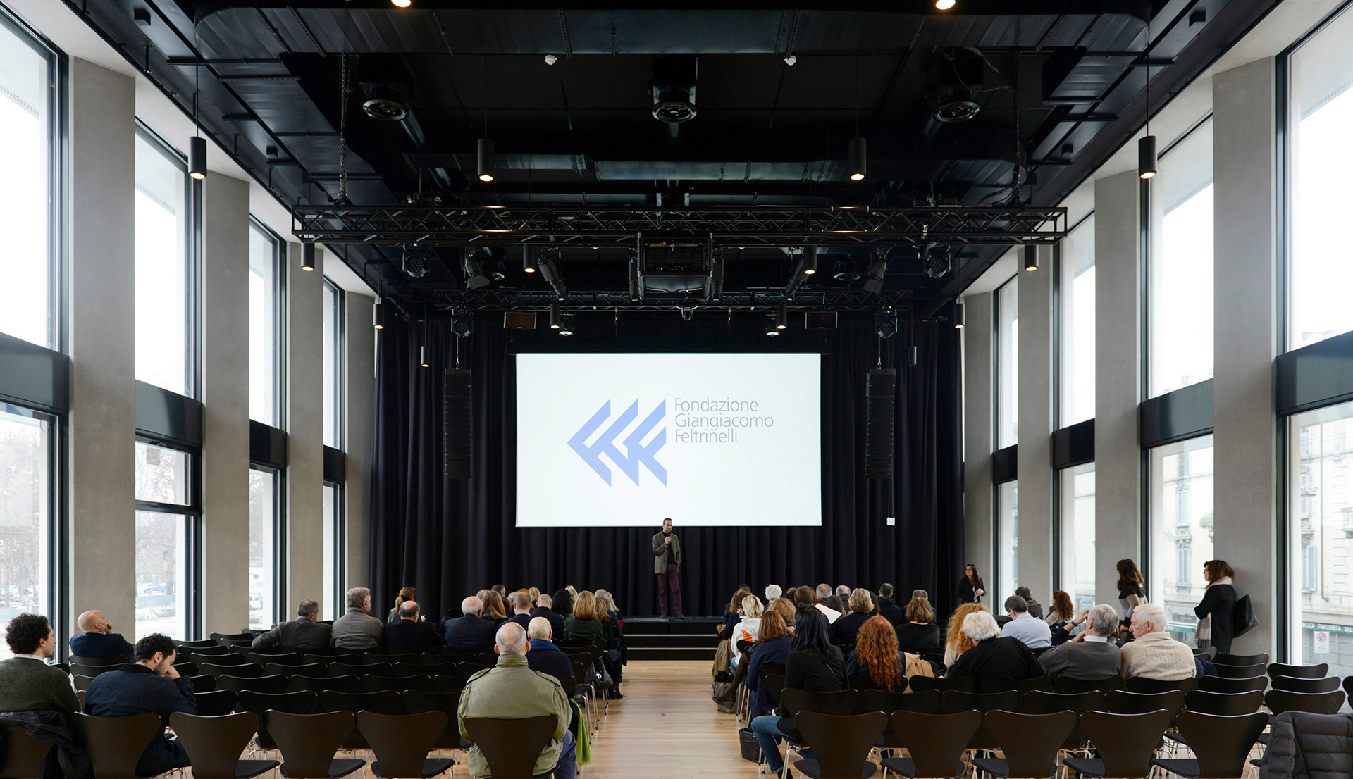 Conference hall with audience seated facing a presenter on stage, large projection screen, tall windows, and industrial ceiling lighting.