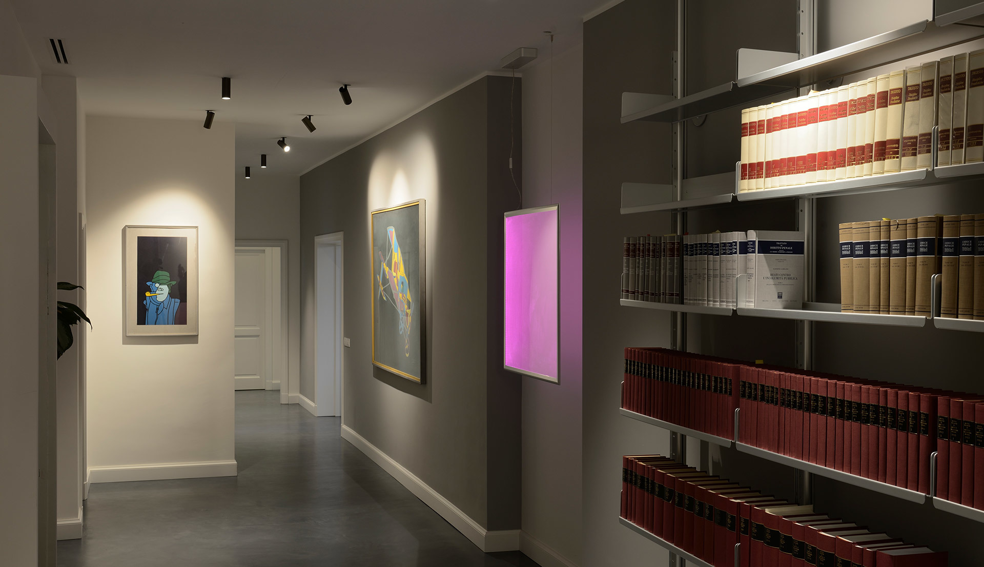 Gallery corridor with track spotlights illuminating framed artworks, a pink glowing wall panel, and shelves of books lit from above
