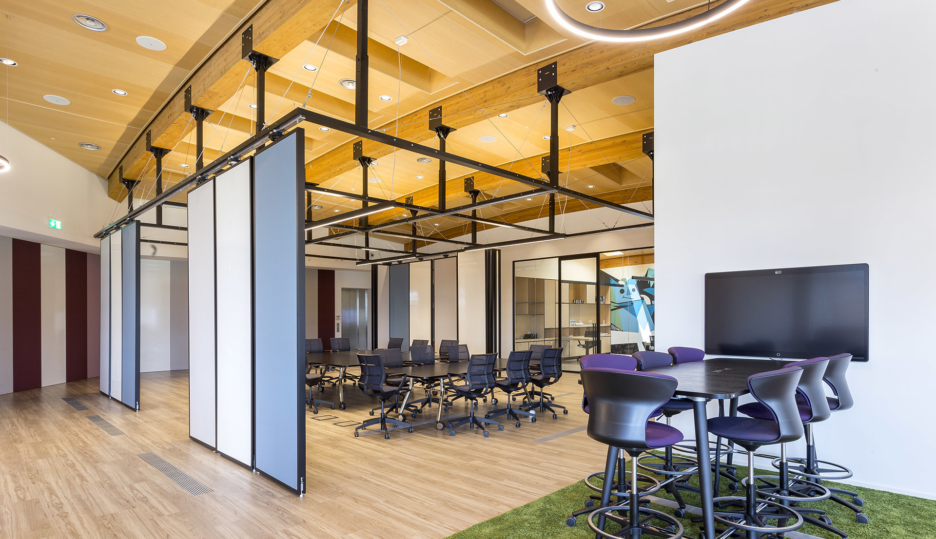 Modern open office with wooden ceiling, black grid track lighting system, movable partitions, conference area, and high bar tables with purple chairs