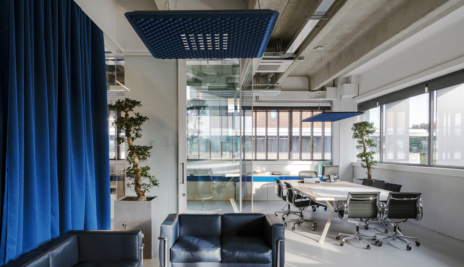 Modern office with blue Artemide pendant lamps over a meeting table, lounge sofa, glass partitions, and bonsai trees.
