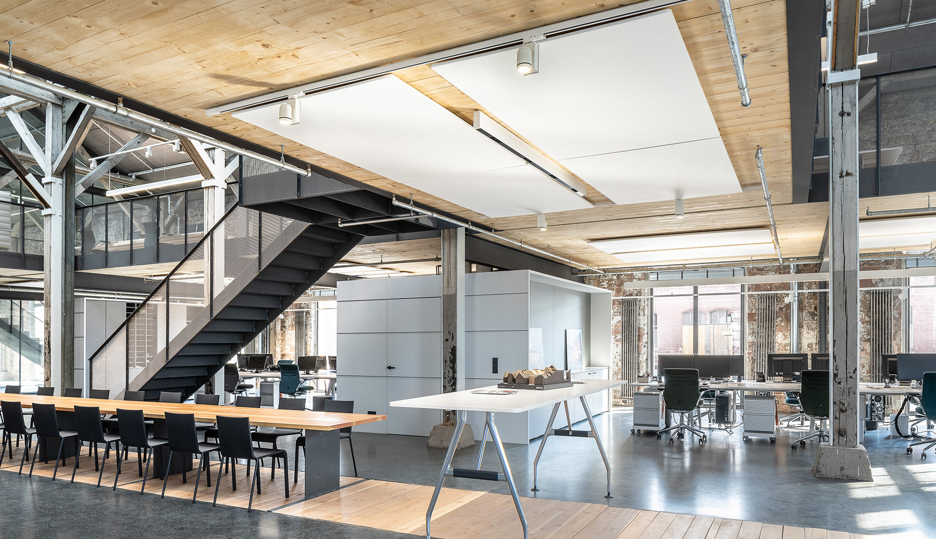Modern open-plan office with Artemide ceiling lights, wooden beams, concrete floors, workstations, and an industrial steel staircase.