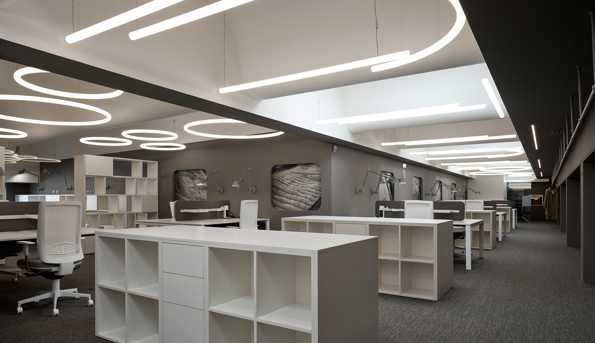 Modern office showroom with Artemide circular pendant lights and linear ceiling fixtures illuminating open workspace with white shelving units.