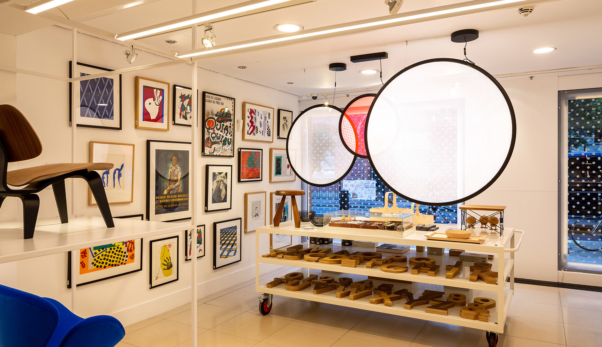 Retail space with large circular pendant lights featuring black frames, a gallery wall of framed art, and a display cart with wooden objects.