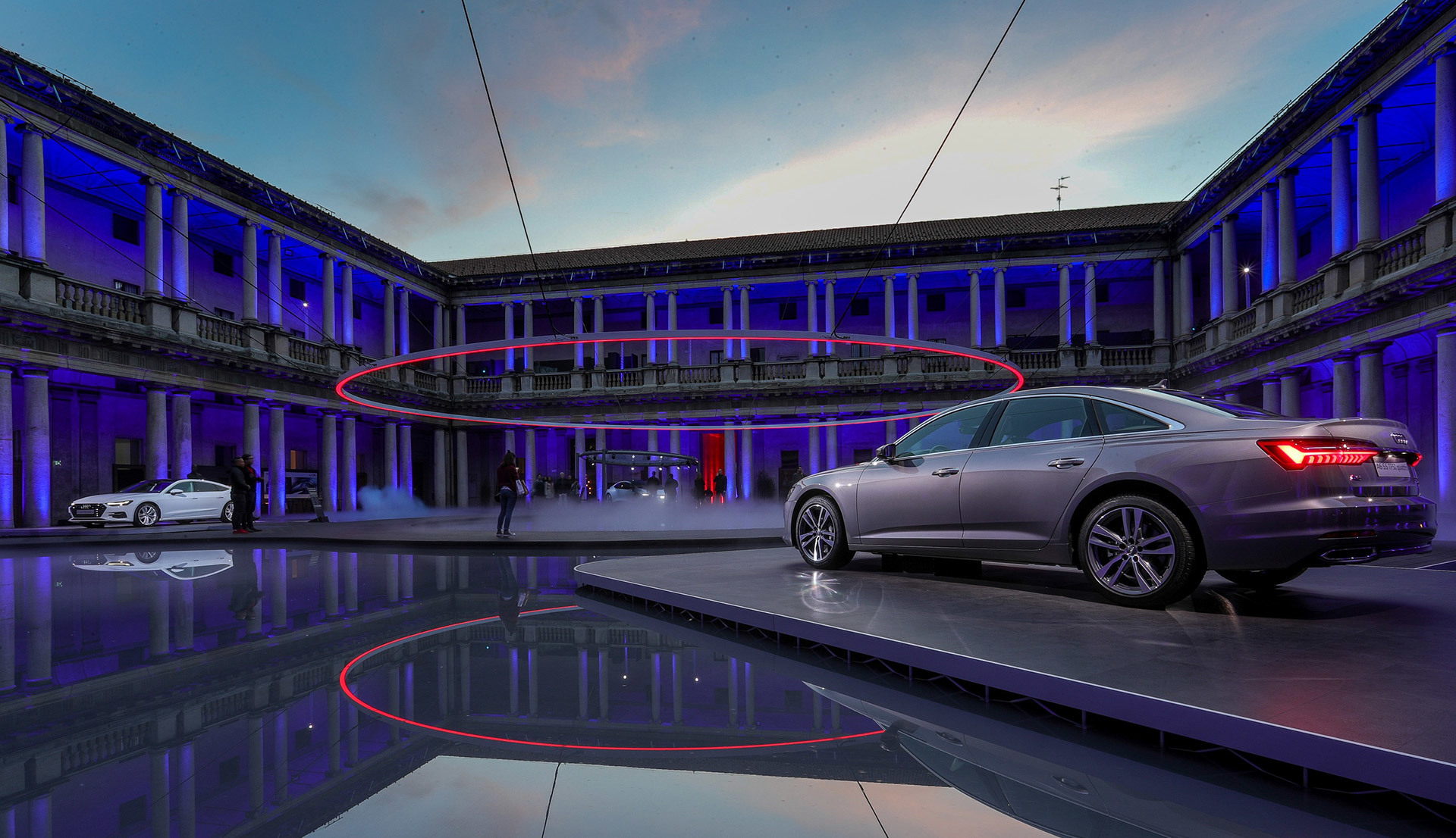 Artemide lighting illuminates a neoclassical courtyard in blue and red, showcasing two luxury cars on a reflective floor at dusk.