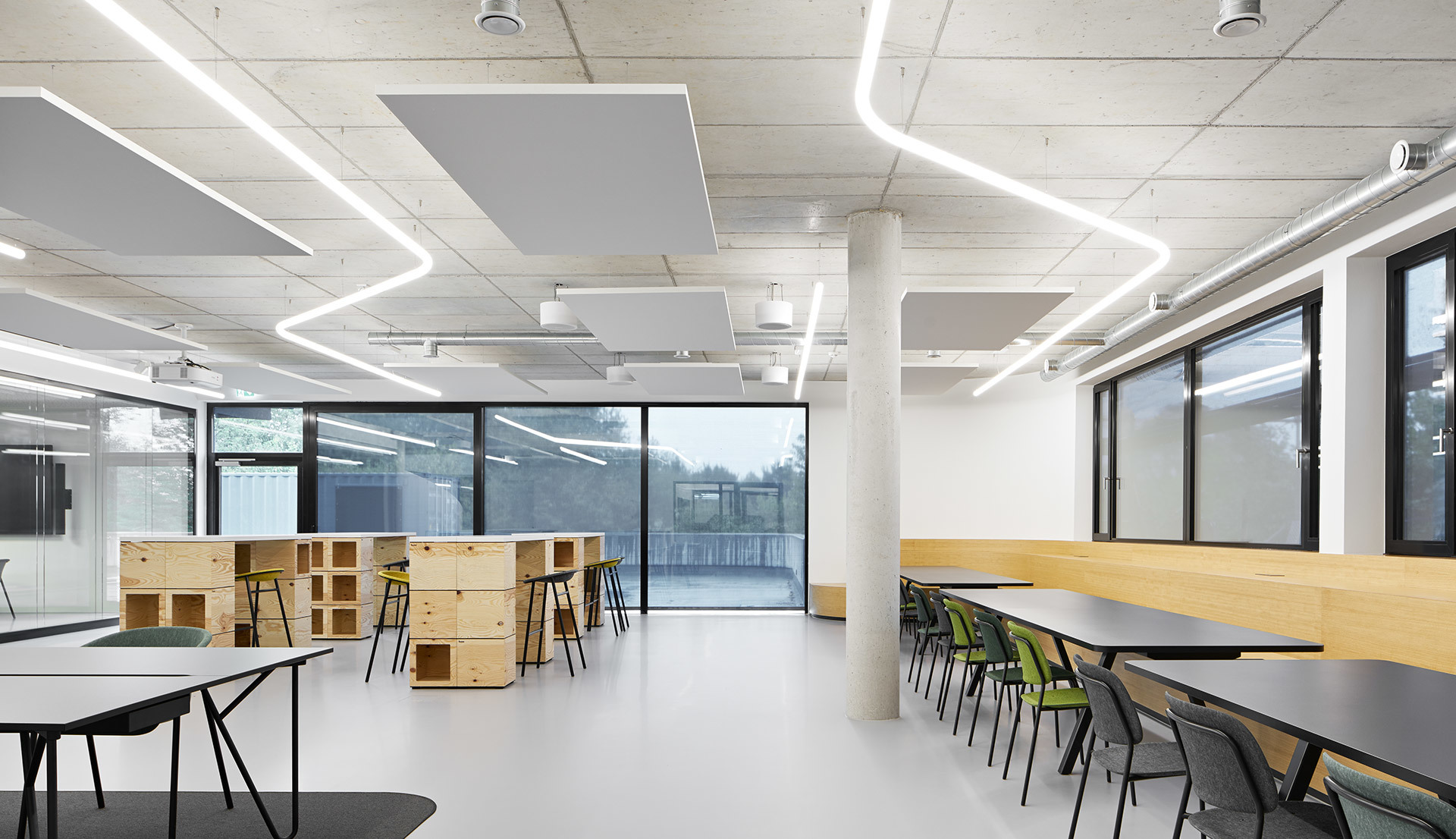 Modern open workspace with geometric linear LED ceiling lights, acoustic panels, wooden modular furniture, and long tables with green chairs.
