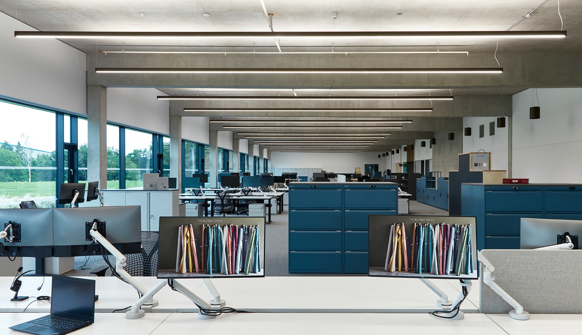 Open-plan office with rows of workstations under linear ceiling-mounted LED light fixtures providing uniform illumination.