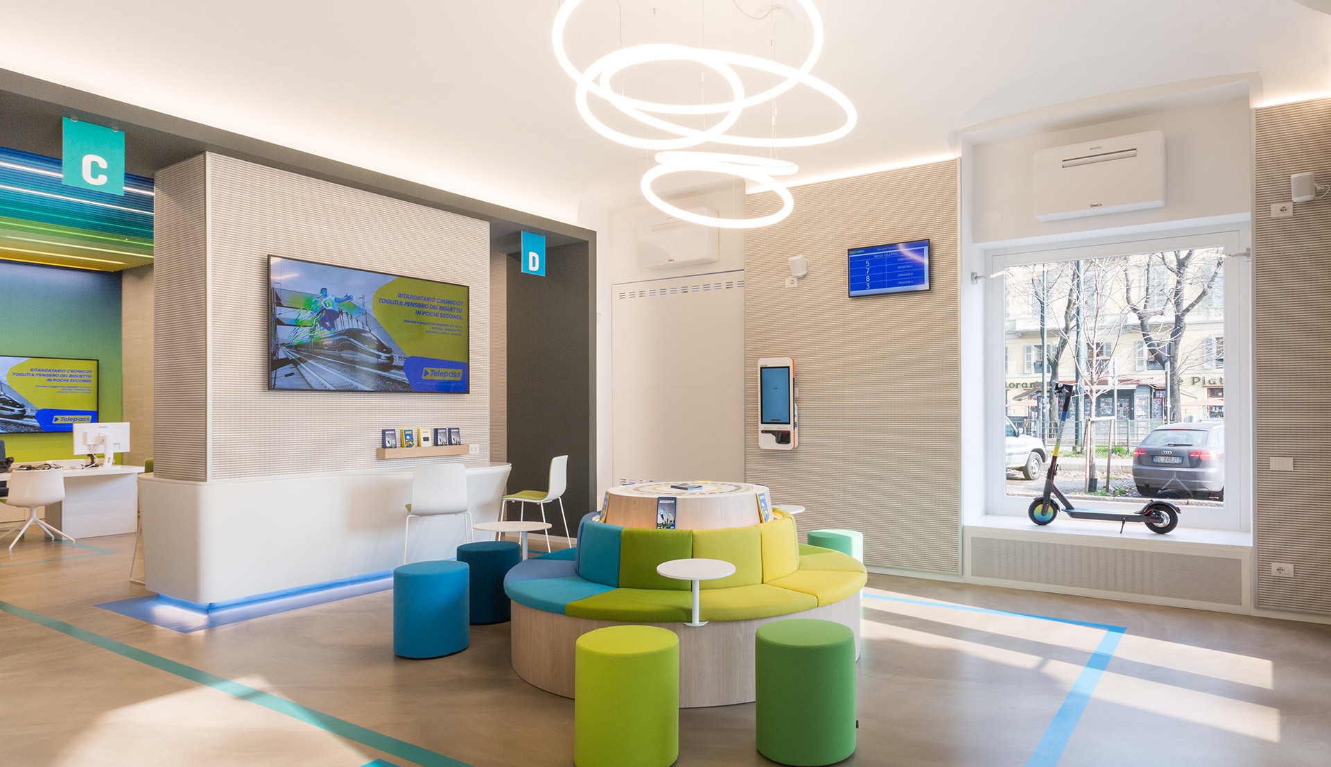 Modern office lounge with Artemide circular ring pendant lights overhead, colorful seating, reception desk, and digital displays.