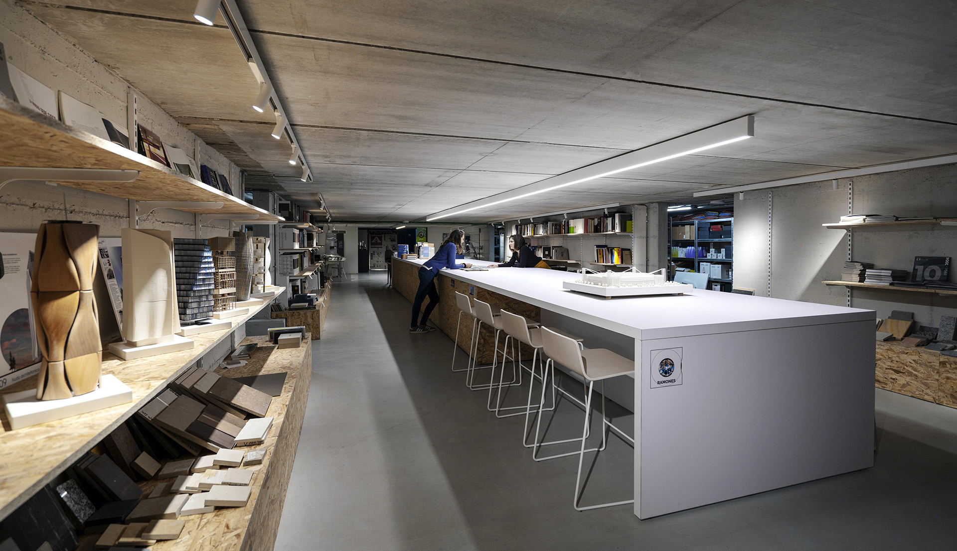 Modern studio workspace with long white table, bar stools, shelving with samples, and Artemide linear LED track lighting on concrete ceiling.