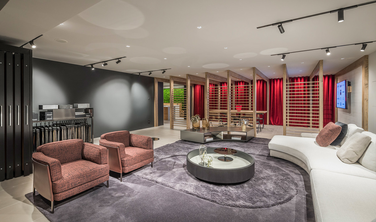 Elegant lounge with track lighting on ceiling, pink armchairs, white curved sofa, round coffee table on grey rug, red curtains backdrop