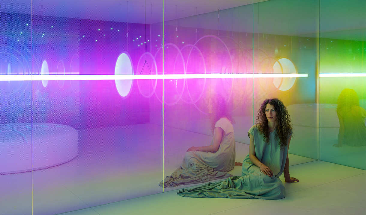 Woman sitting in an immersive room bathed in vibrant pink, purple, and green light from a horizontal neon tube installation