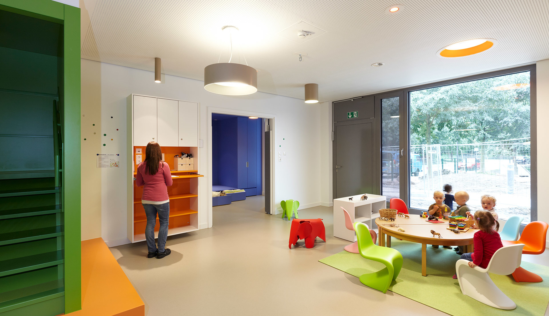 Bright children's playroom with colorful furniture, pendant ceiling lamp, and recessed lighting illuminating kids at a round table.
