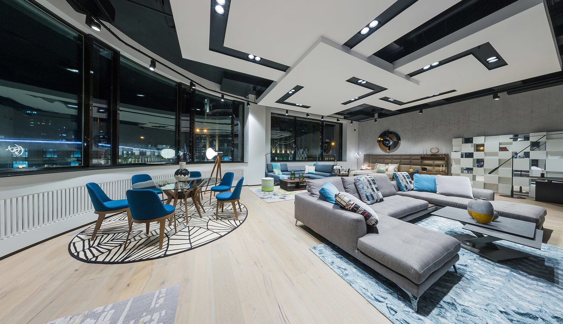 Modern open-plan living space with recessed ceiling lights, large gray sectional sofa, blue dining chairs, and floor-to-ceiling windows at night
