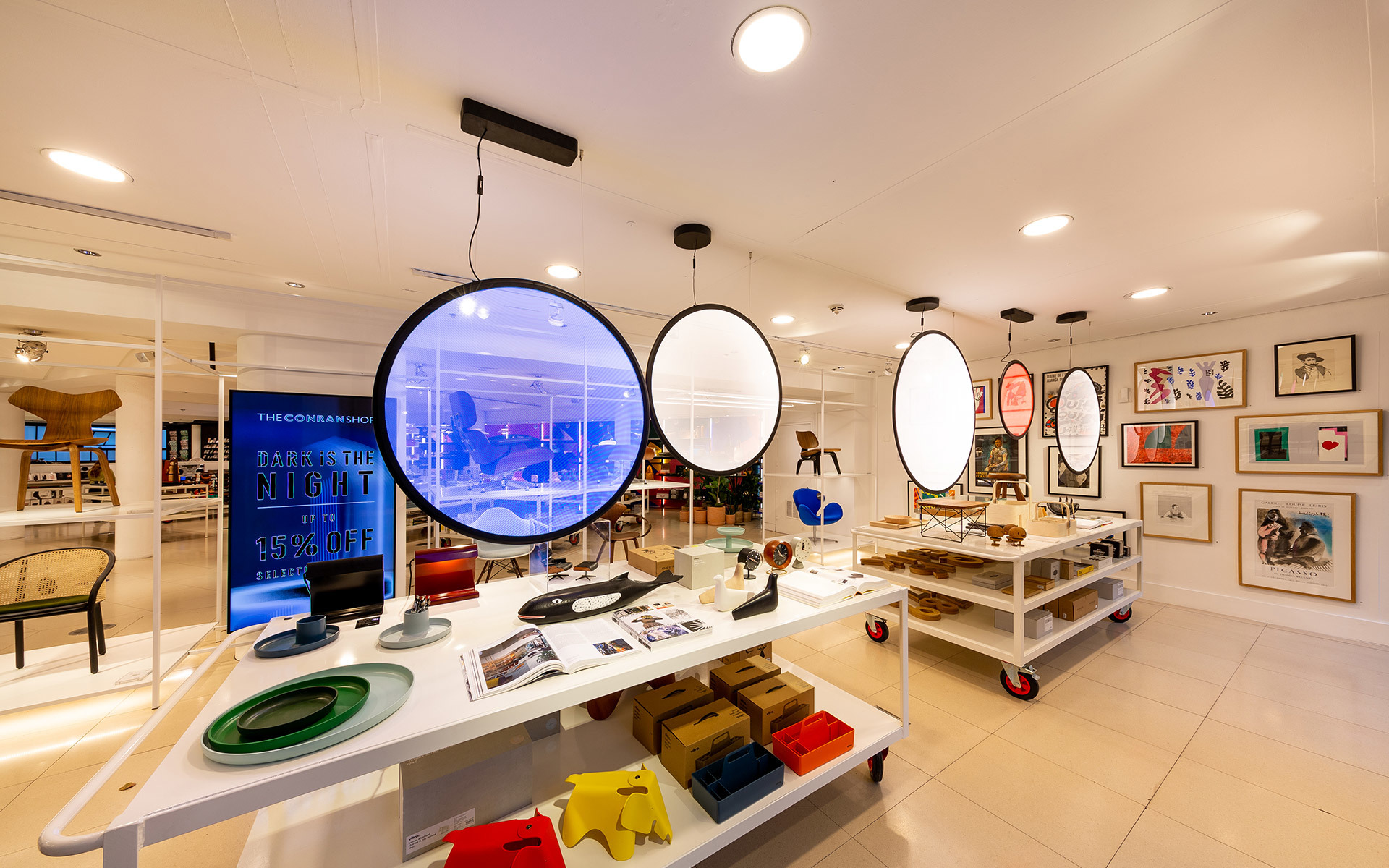 Modern retail space with large circular pendant lights illuminating display tables featuring colorful design objects and accessories.