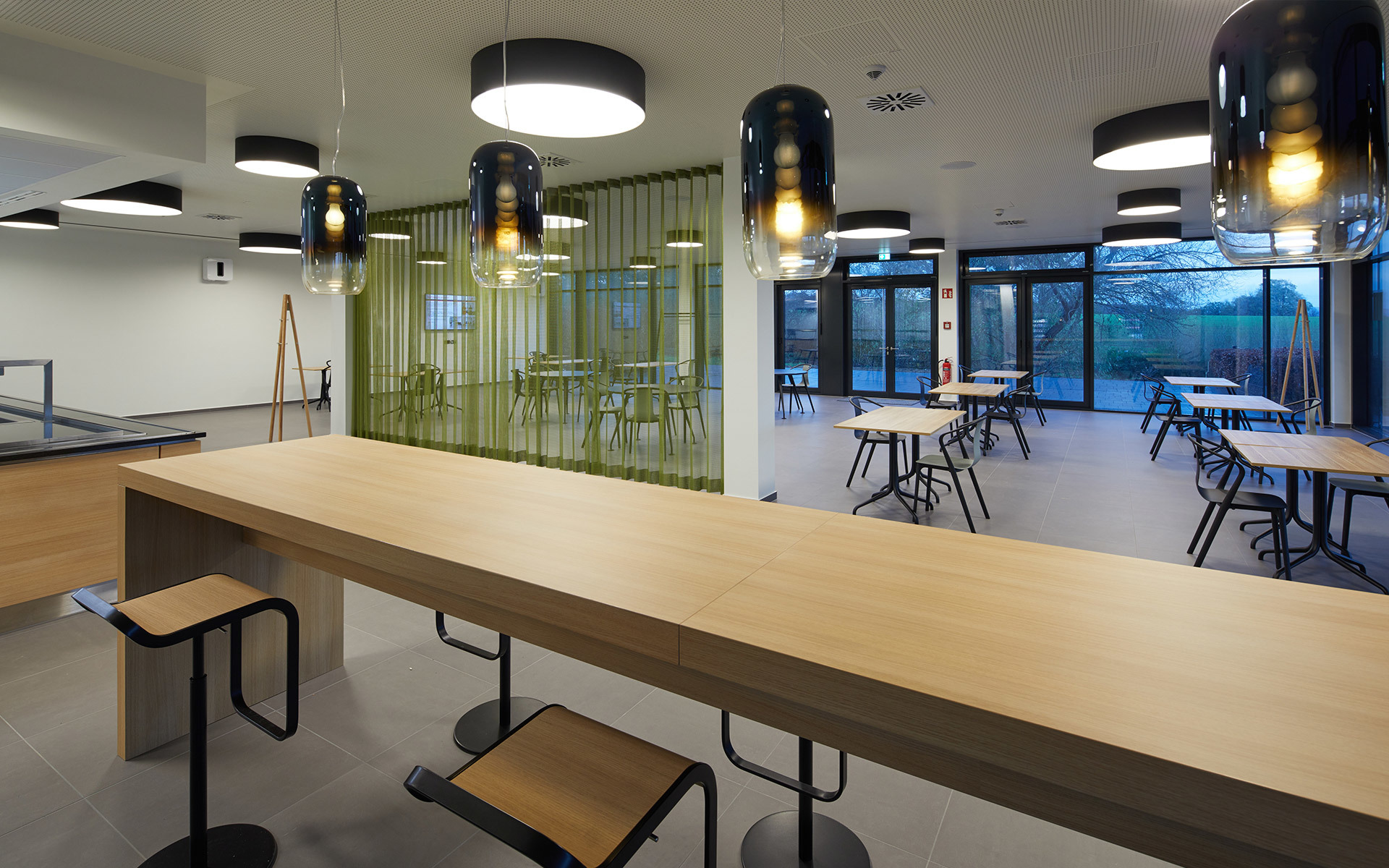 Modern cafeteria with Artemide pendant lamps in smoked glass over a long wooden bar counter and small tables with black stools