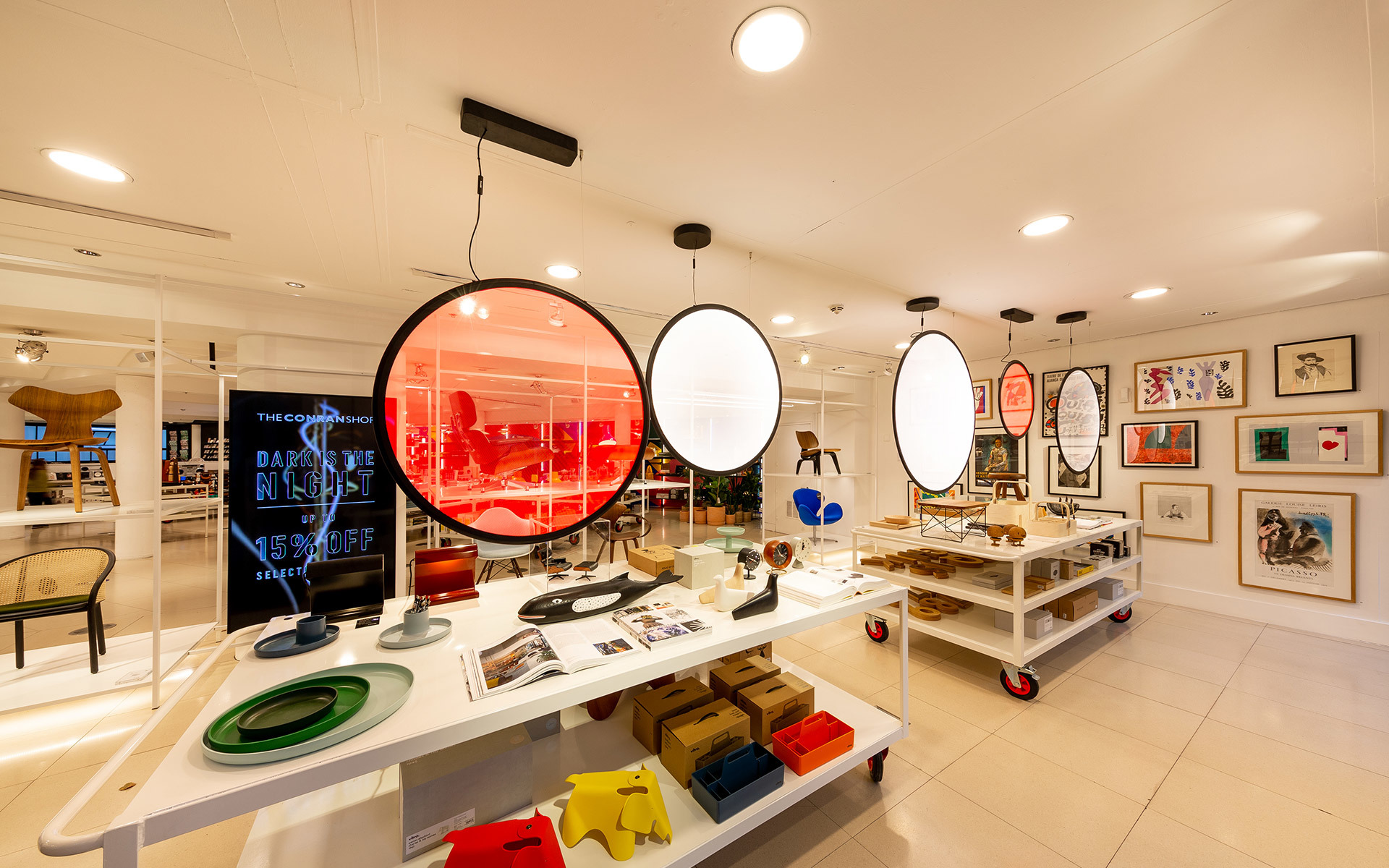 Modern design shop with large oval pendant lights, white display tables showcasing colorful objects, and framed artwork on walls.