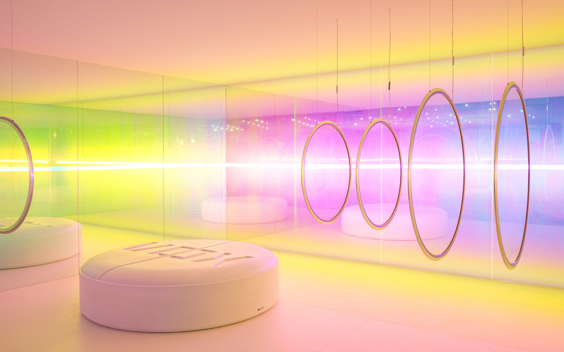 Immersive room with circular pendant ring lights casting vibrant rainbow gradients across mirrored walls and white curved seating