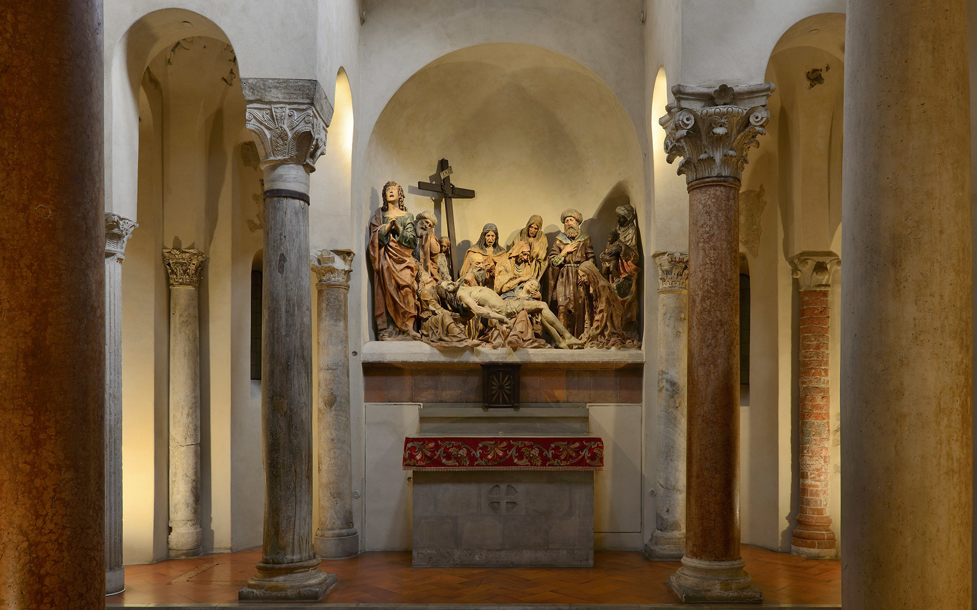 Chapel interior with stone columns, arched niches, and a polychrome sculptural Pietà group illuminated by warm accent lighting above an altar.