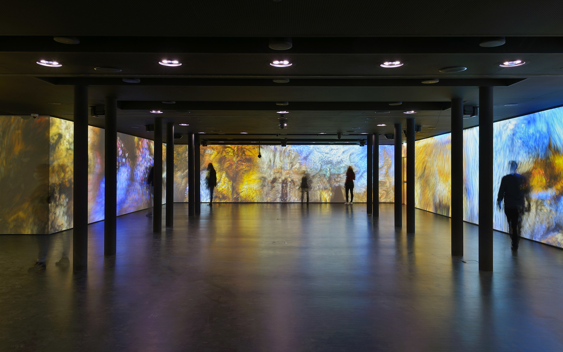 Immersive art installation in a dark hall with colorful projected murals in blue and gold, visitors silhouetted, ceiling spotlights above.
