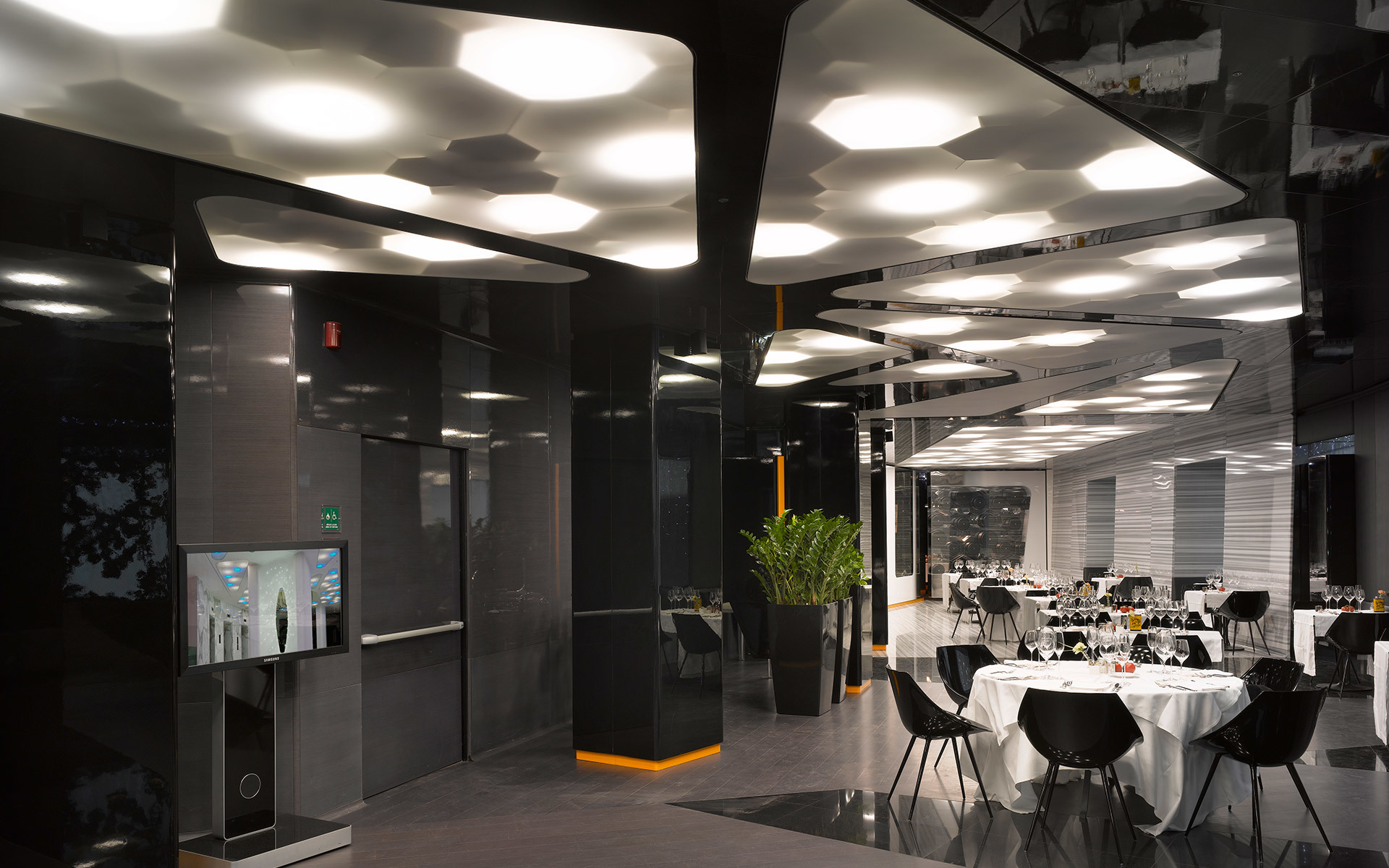 Modern restaurant interior with hexagonal ceiling lights illuminating dining tables with white tablecloths in a dark, sleek space