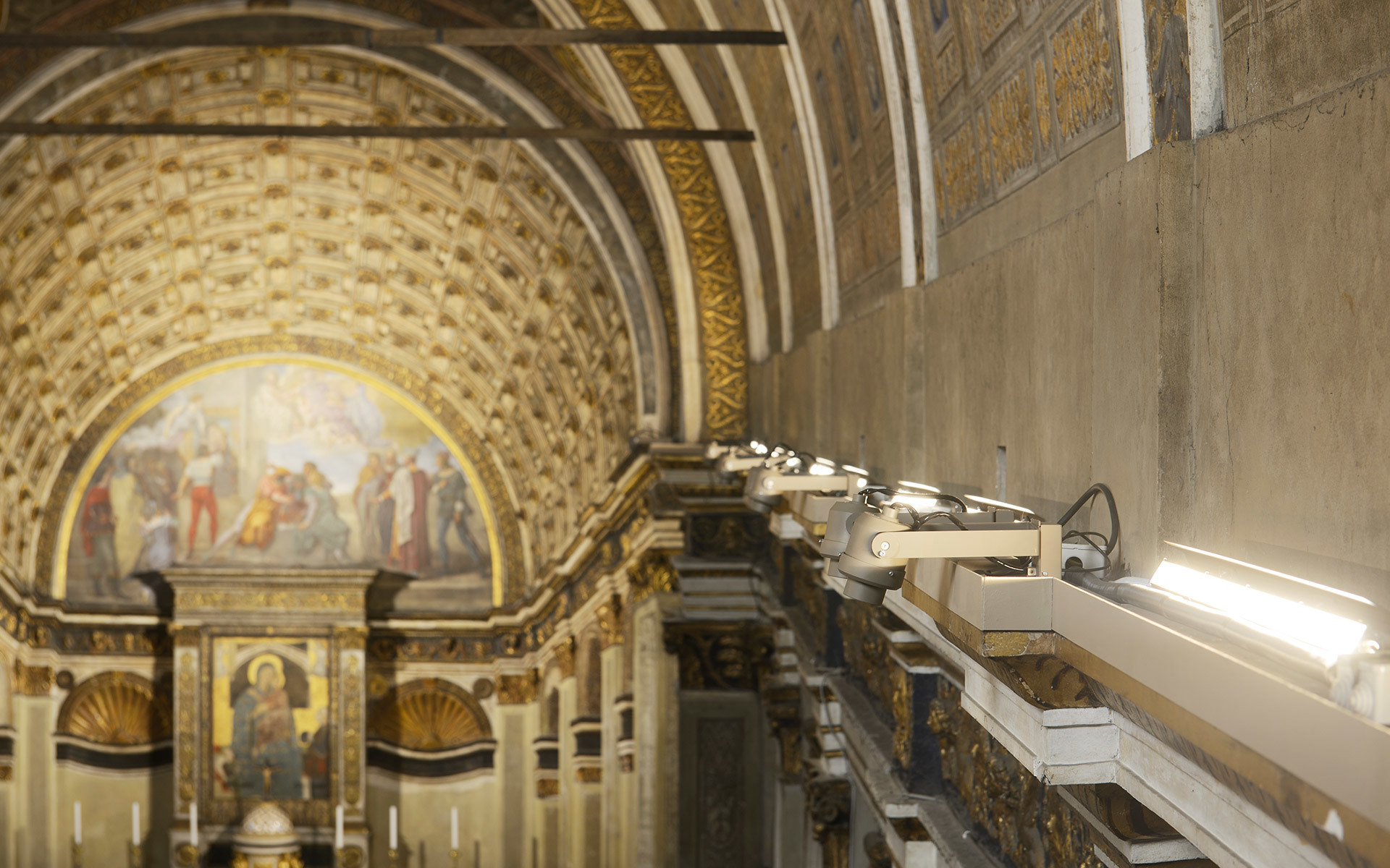 Artemide lighting fixtures mounted on a rail illuminate the ornate frescoed apse and gilded altar of a historic church.