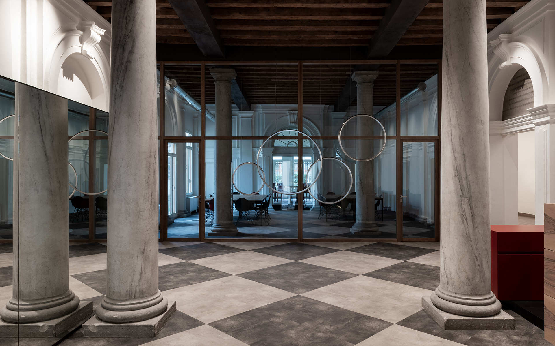 Elegant hall with marble columns and checkered floor, featuring circular ring pendant lights suspended behind glass partitions.