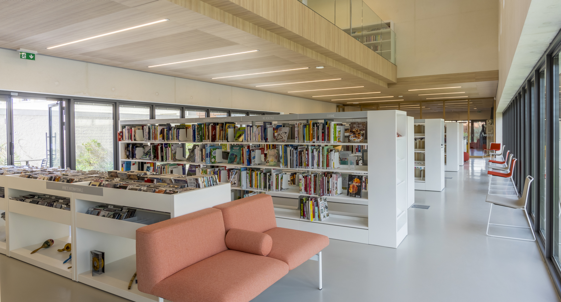 Modern library interior with Artemide linear ceiling lights illuminating open bookshelves, a pink sofa, and large windows.