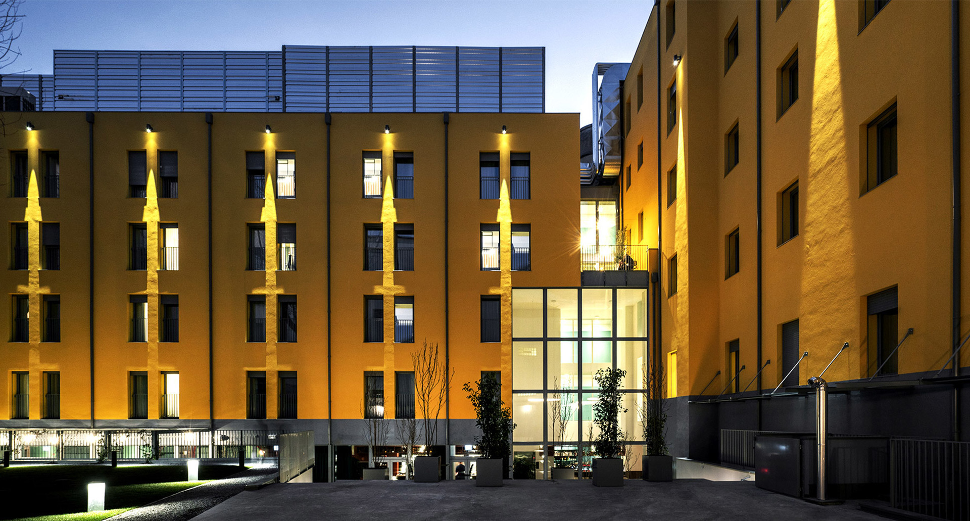 Modern building facade at dusk, illuminated by warm yellow wall-mounted uplights and bollard lights in the courtyard