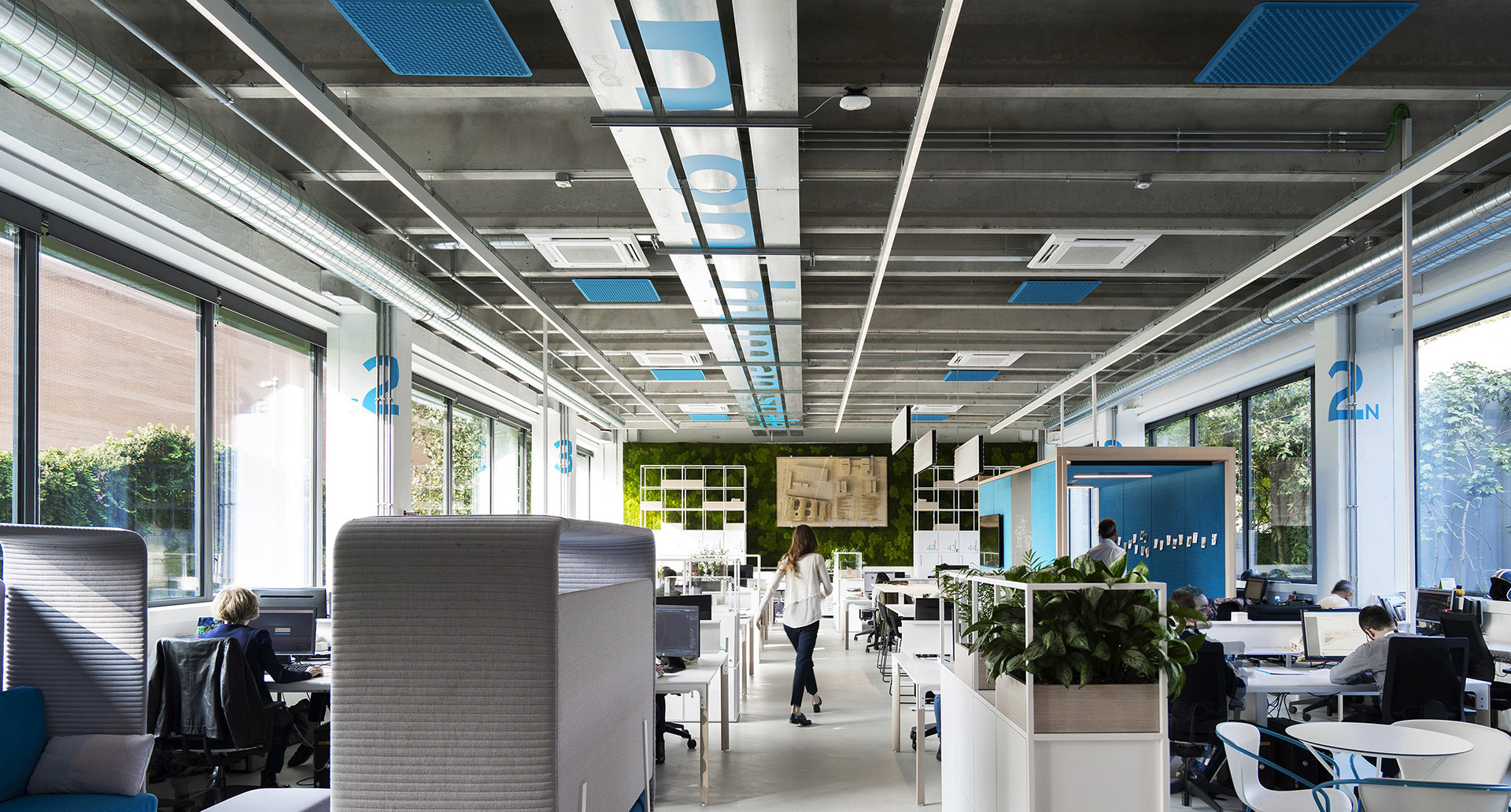 Modern open-plan office with Artemide ceiling lighting panels, workstations, acoustic pods, and green plants under natural light.