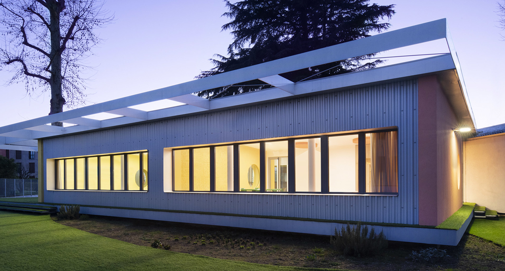 Modern single-story building exterior at dusk, illuminated by warm interior lighting through large windows and skylights.