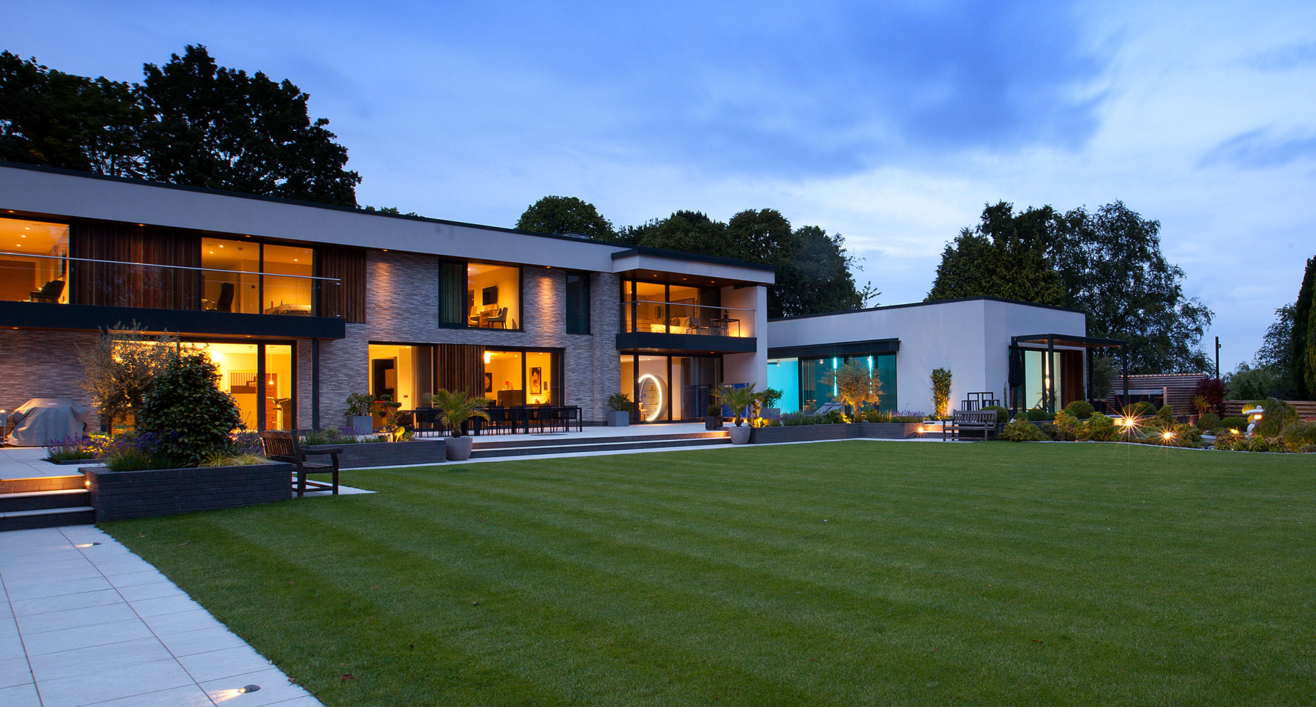 Modern two-story villa at dusk with warm interior lighting and subtle outdoor ground lights illuminating the landscaped lawn
