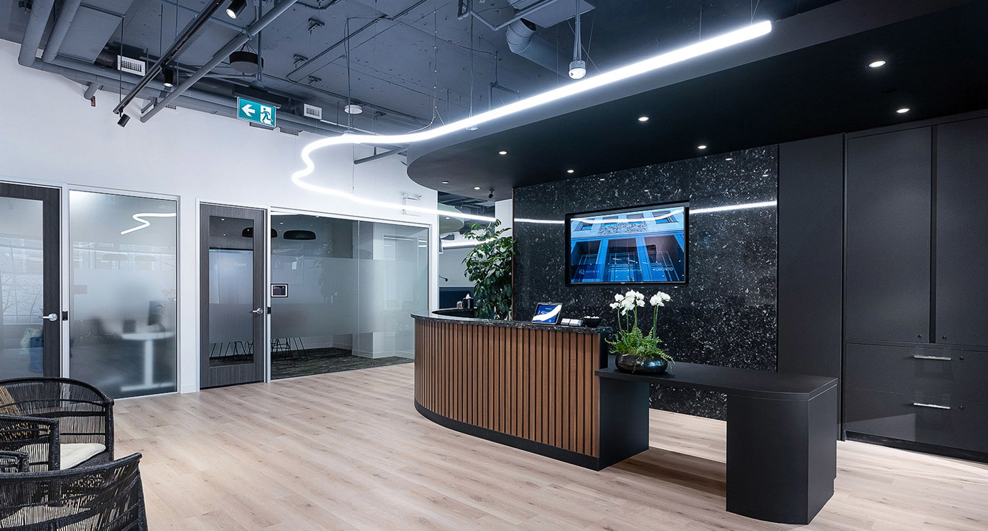 Modern office reception with curved Artemide LED linear light on dark ceiling, wooden desk, marble wall, and glass meeting rooms.