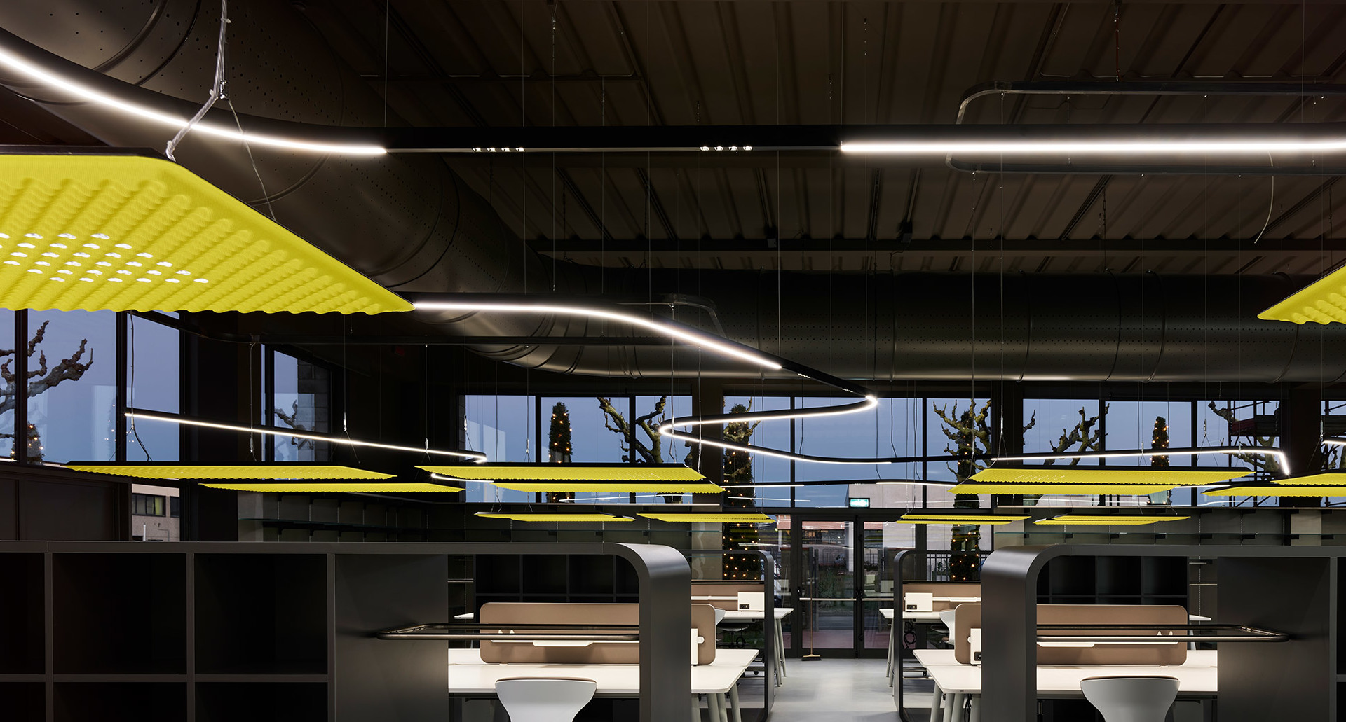 Modern office with curved Artemide pendant lights and yellow acoustic panels suspended from dark industrial ceiling above workstations