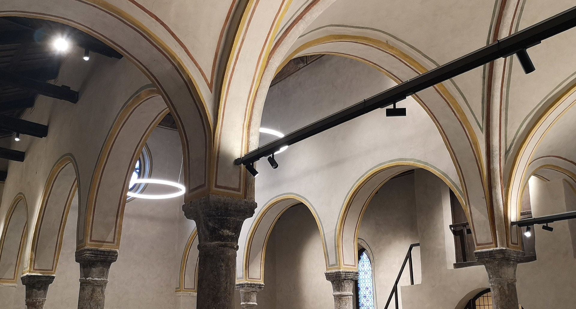 Gothic vaulted hall with Artemide circular LED ring pendant and black track spotlights illuminating stone arches and columns