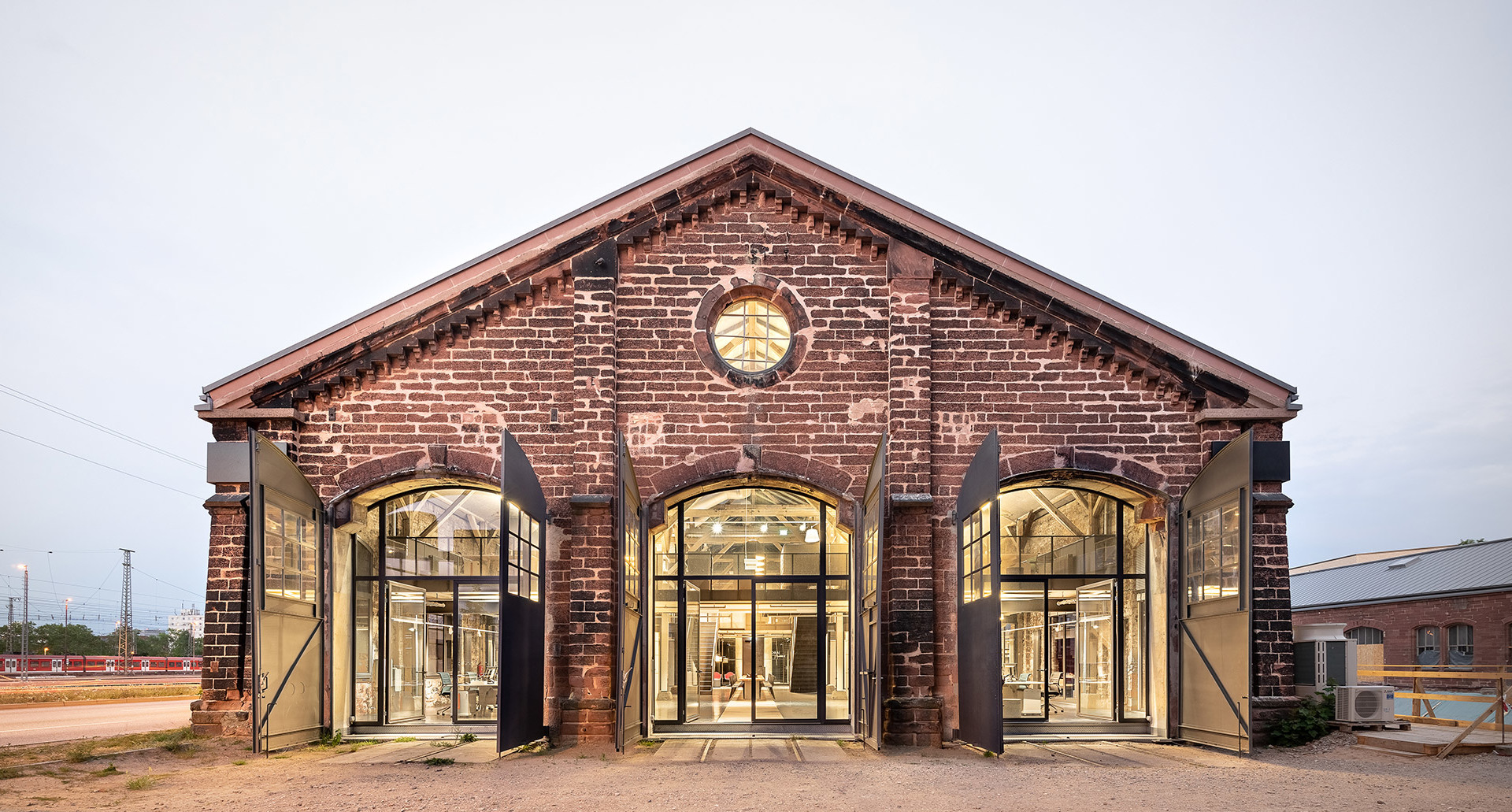 Renovated historic red brick industrial building with large arched windows, warmly lit interior visible at dusk.