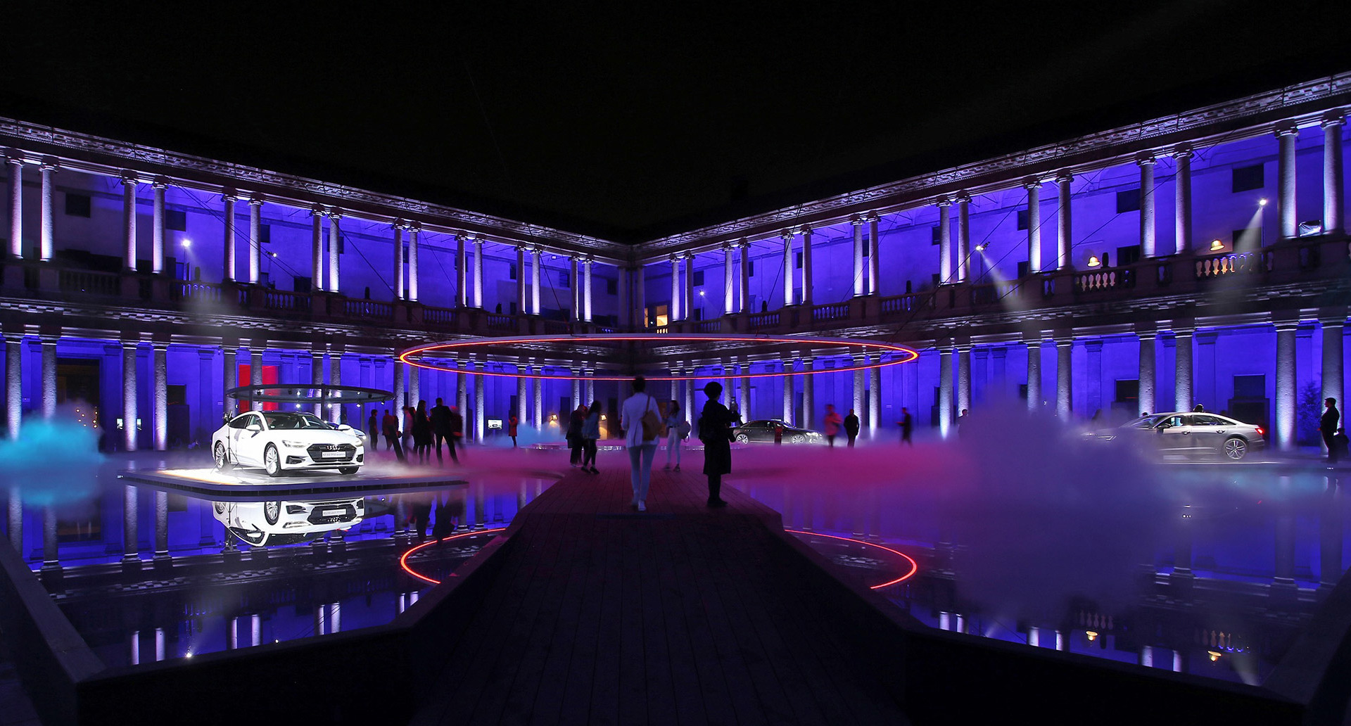 Grand neoclassical courtyard lit in deep blue with illuminated columns, two white cars on display, red circular light installation, and fog effects at night