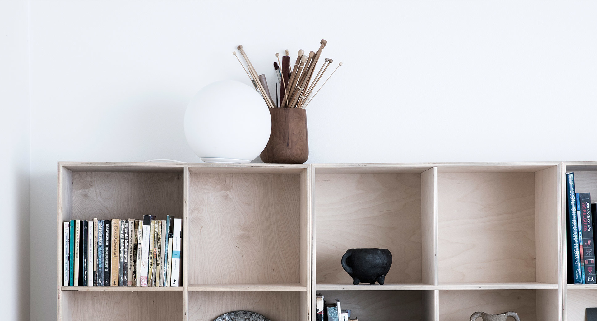 Light wood modular bookshelf with books and decorative objects, topped with a round white globe lamp and a vase with sticks