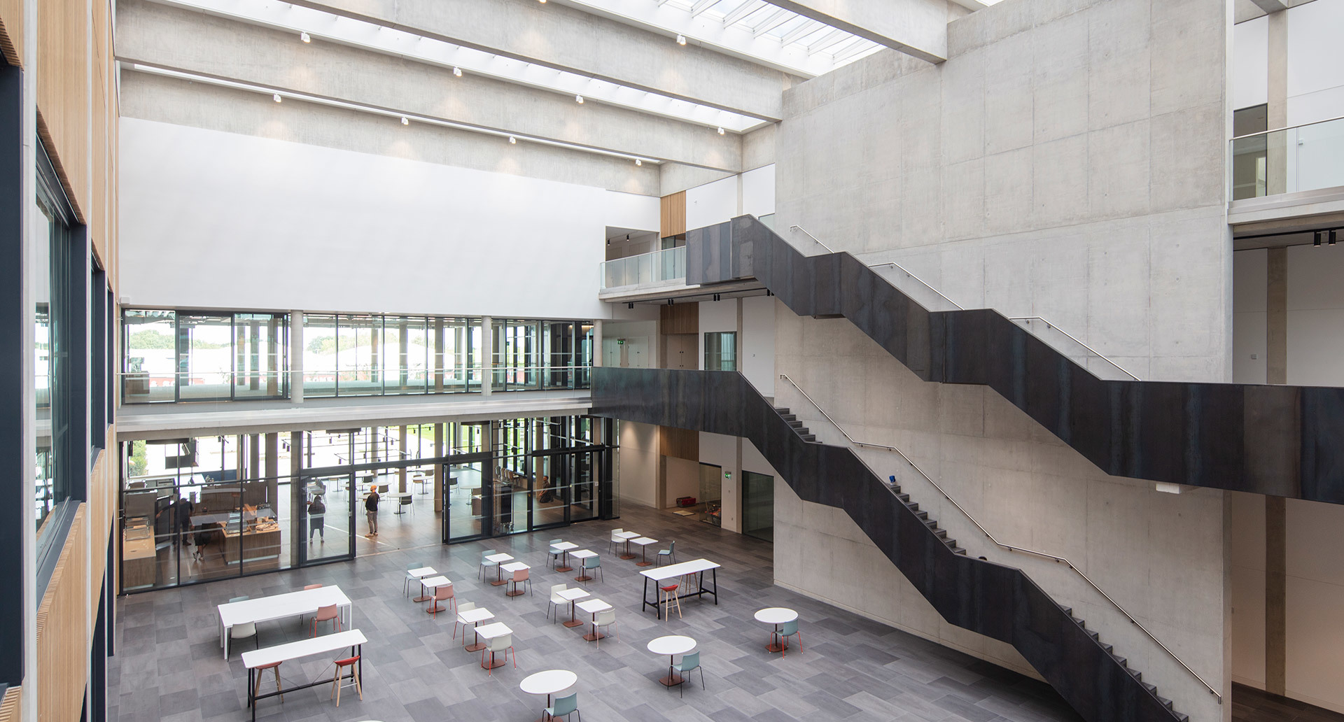 Modern atrium with skylights, linear ceiling fixtures, white tables, stools, concrete walls, and a dark metal staircase on two levels.