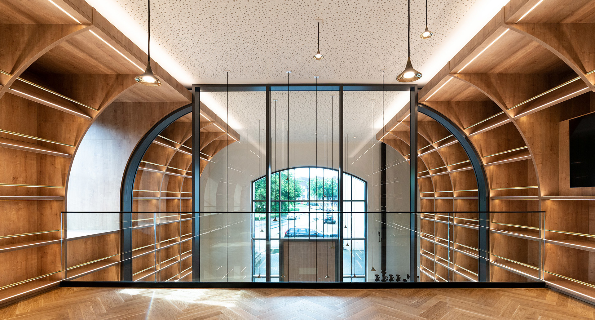 Elegant interior with warm wood shelving, arched glass partition, herringbone floor, and Artemide pendant lights hanging from a perforated ceiling.