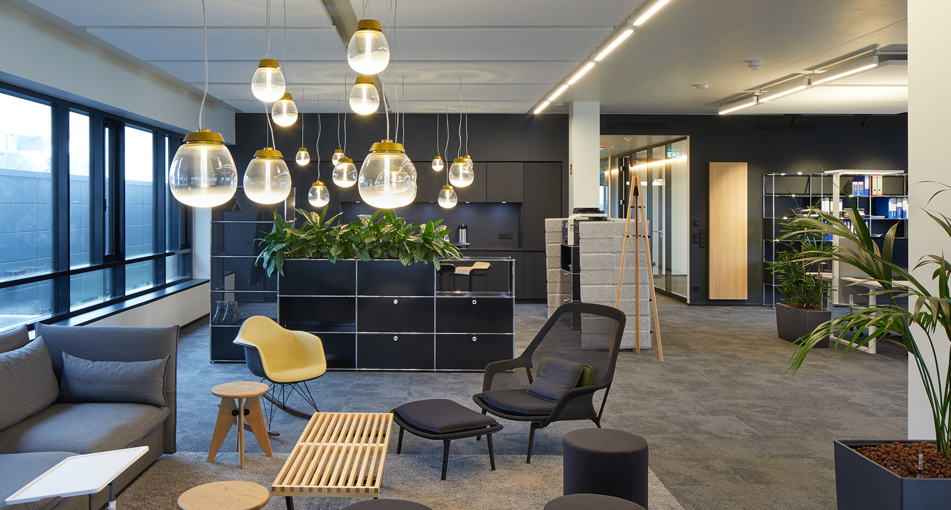Modern office lounge with Artemide pendant lights featuring gold-topped glass globes, grey sofas, and lush green plants.
