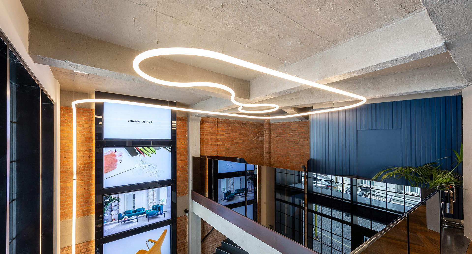 Artemide custom freeform LED light installation suspended from concrete ceiling over a modern industrial mezzanine with brick walls.