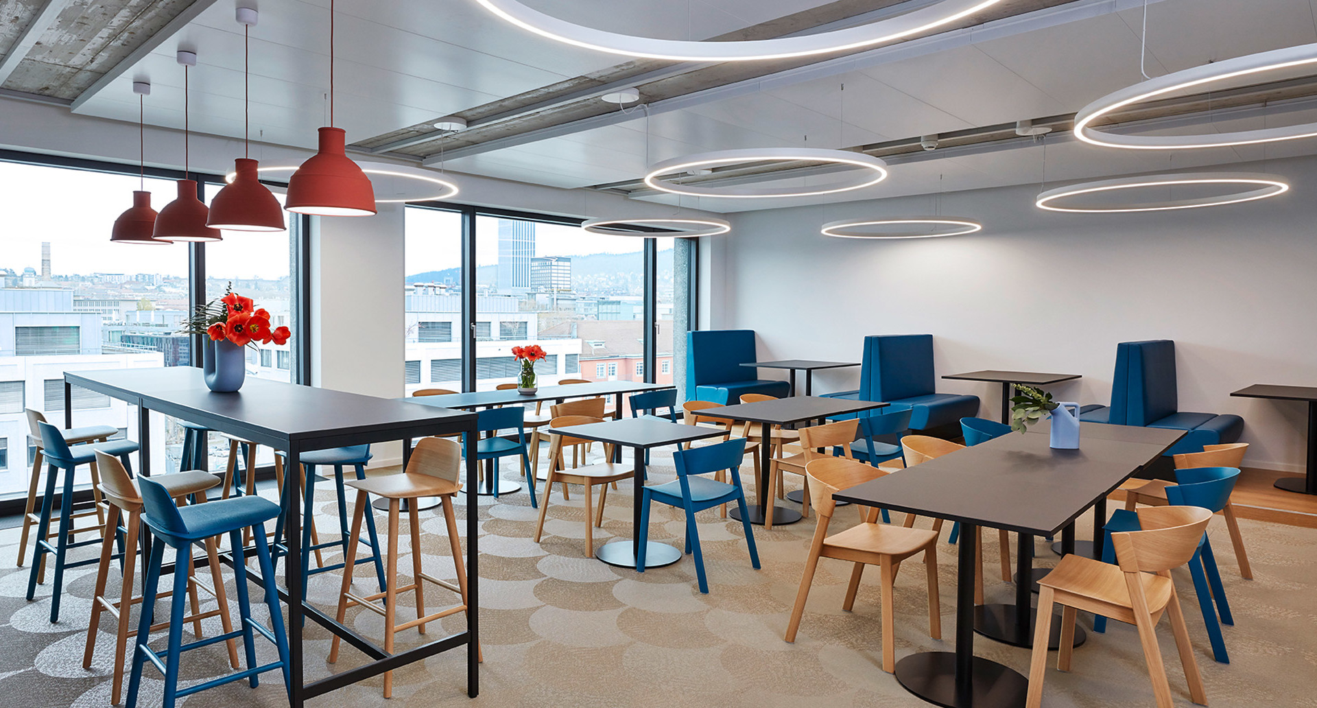 Modern office cafeteria with Artemide circular LED ceiling rings, red pendant lamps, blue and wood chairs, large windows with city view