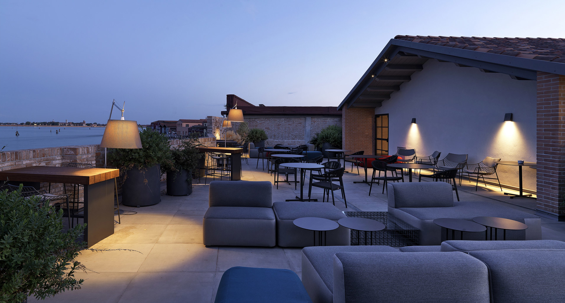 Rooftop terrace at dusk with Artemide floor and wall lamps illuminating lounge seating and dining areas overlooking the water